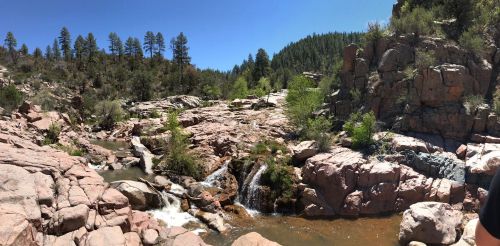 A scenic view of a rocky riverbank with a small waterfall, surrounded by trees and blue sky.