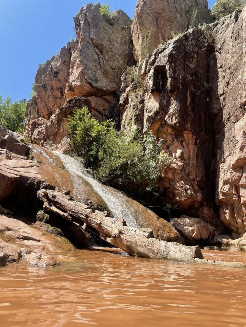 A rocky landscape with a waterfall cascading into a brown pool, surrounded by lush greenery and steep cliffs.