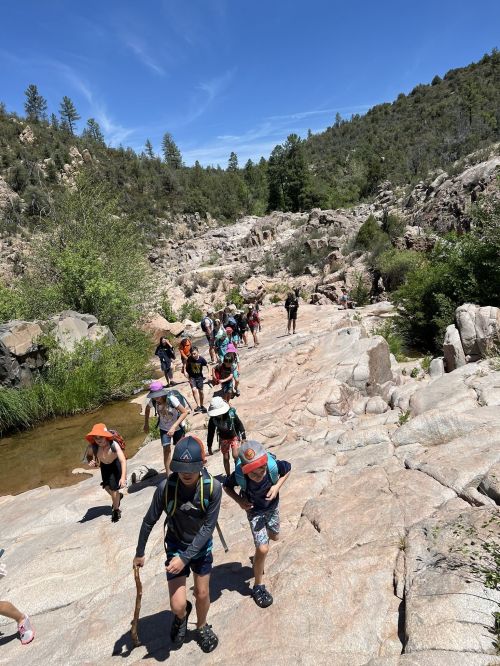 A group of children hiking along rocky terrain near a stream, surrounded by trees and mountains under a clear blue sky.