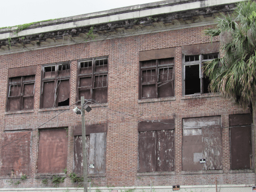 Abandoned brick building with boarded-up windows and overgrown vegetation.