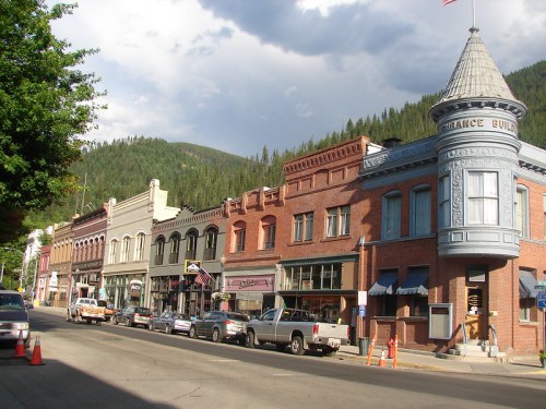Historic buildings line a street in a mountain town, featuring a mix of architectural styles and greenery in the background.