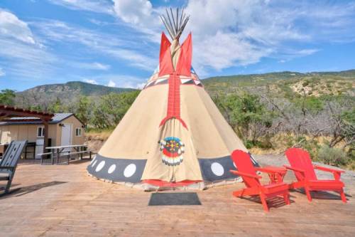 A colorful teepee stands on a wooden deck, surrounded by mountains and trees, with red chairs in front.