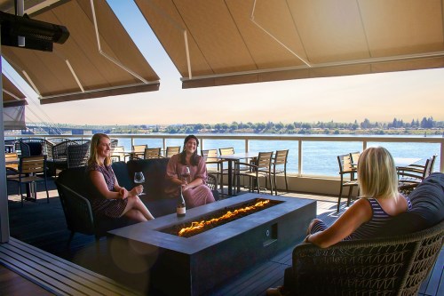 Three women relax on a patio by a river, enjoying drinks around a fire pit with scenic views in the background.