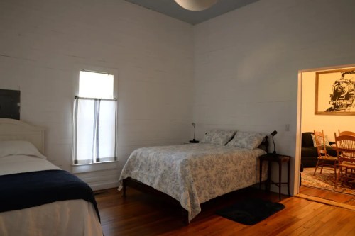 A cozy bedroom with two beds, a window, and an open door leading to a living area with a dining table.