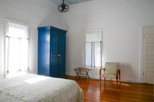 A bright, minimalist bedroom with a blue wardrobe, wooden floors, and a simple chair beside a small table.