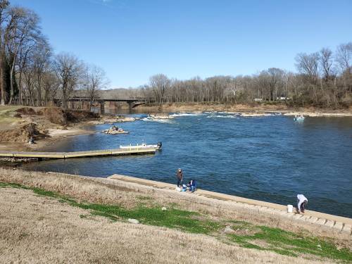 A river scene with a dock, people fishing, and a bridge in the background under a clear blue sky.