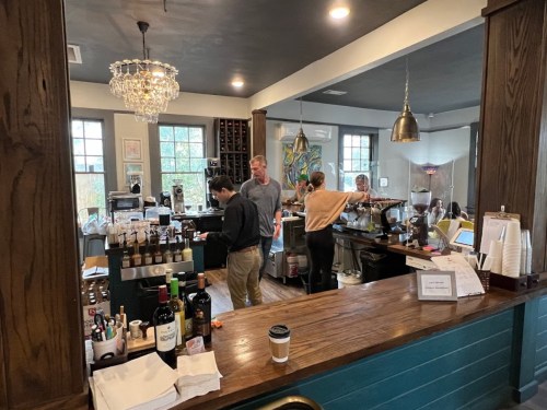 A cozy café interior with a barista serving customers, wine bottles on display, and a chandelier overhead.
