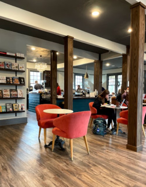 Cozy café interior with wooden floors, red chairs, and patrons working at tables. Bookshelves line the wall.