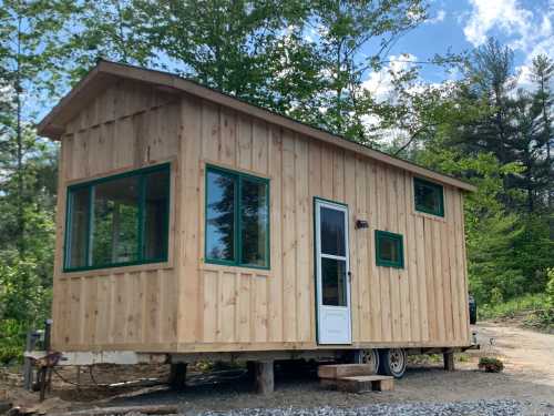 A small wooden cabin on wheels, featuring large windows and a white door, surrounded by trees and blue sky.