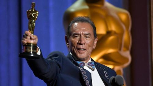 A man in a traditional outfit holds an Oscar trophy, smiling proudly on stage with a golden statue in the background.