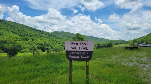 Sign for West Fork Rail Trail in Monongahela National Forest, surrounded by lush green hills and a blue sky with clouds.