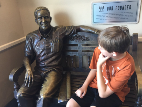 A boy sits next to a bronze statue of a smiling man on a bench, with a plaque titled "Our Founder" in the background.