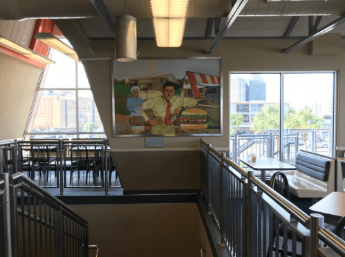 Interior of a restaurant featuring a large mural of a man with burgers, large windows, and a staircase.