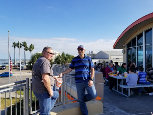 Two men stand outside a restaurant with a view of palm trees and a marina, enjoying a sunny day.