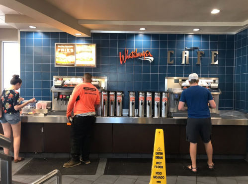 Two customers at a fast-food counter with drink dispensers, a caution sign on the floor, and menu boards above.