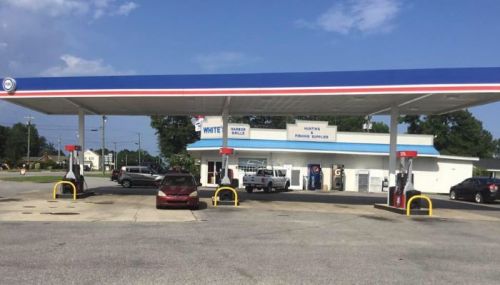 A gas station with multiple pumps, cars parked, and a convenience store in the background under a blue sky.
