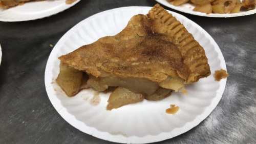 A slice of apple pie on a paper plate, showcasing a flaky crust and visible apple filling.