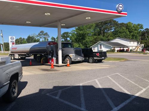A fuel tanker truck refuels at a gas station, with a pickup truck parked nearby and trees in the background.