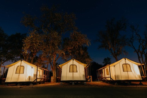 Three glamping tents illuminated at night, surrounded by trees under a starry sky.