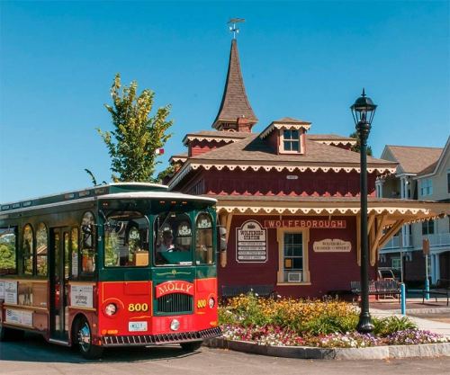 A colorful trolley in front of a charming red train station with a tower and flower beds in a sunny setting.