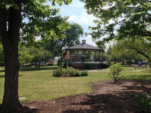 A gazebo in a park surrounded by trees and greenery, with a clear blue sky in the background.