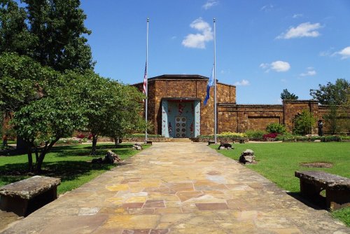 A stone building with blue doors, surrounded by green grass and trees, under a clear blue sky. Flags are at half-mast.