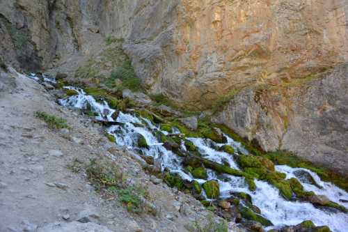 A serene stream flows over rocks, surrounded by moss and steep rocky cliffs in a natural landscape.