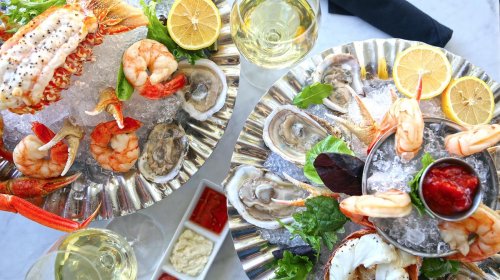 Two seafood platters on ice, featuring shrimp, lobster, oysters, lemon slices, and dipping sauces, with glasses of white wine.