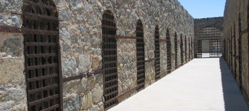 A stone corridor with barred windows and doors, leading to a bright, open space at the end.