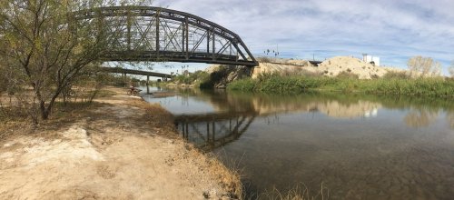 A riverbank scene featuring a bridge overhead, with greenery and a calm water reflection under a cloudy sky.