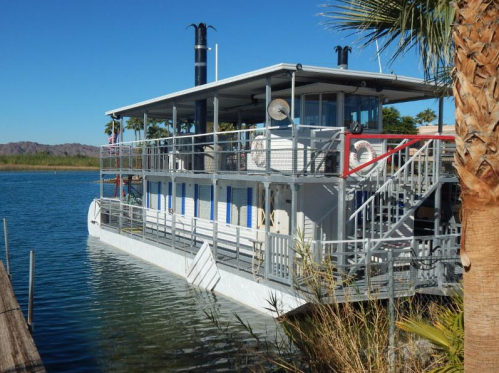 A large houseboat docked by the shore, surrounded by palm trees and calm water under a clear blue sky.
