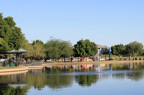A serene park scene with a pond, trees, and playground equipment in the background under a clear blue sky.