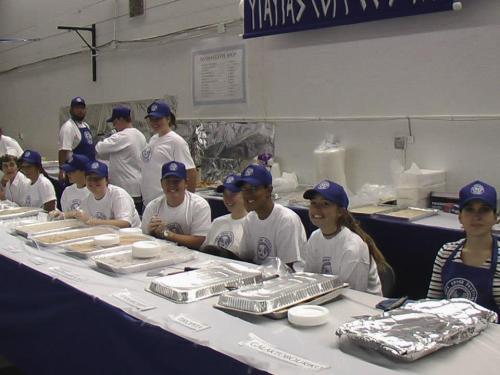 A group of volunteers in blue hats and white shirts smiles behind a table with food trays at a community event.
