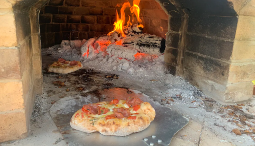 A wood-fired oven with two pizzas baking on a metal peel, surrounded by glowing embers and ash.