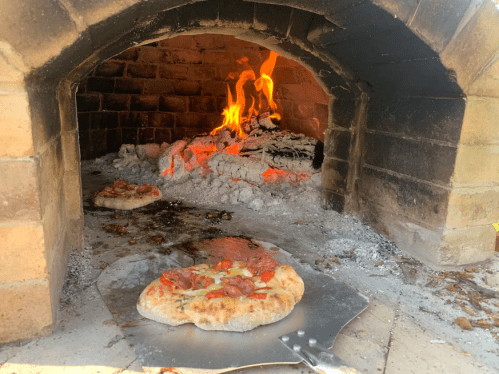 A wood-fired oven with flames, featuring a pizza on a metal peel in front of it.