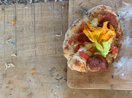 A rustic pizza topped with sausage, tomatoes, cheese, and a yellow flower, resting on a wooden cutting board.