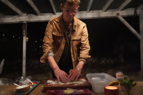 A person in a brown jacket prepares food on a wooden table outdoors, with candles and ingredients around.