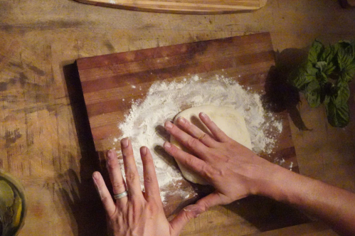 Hands kneading dough on a floured wooden surface, with fresh mint leaves nearby.