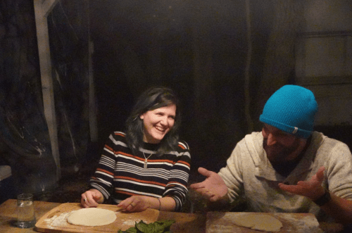 Two people sitting at a table, laughing and preparing dough in a dimly lit room.