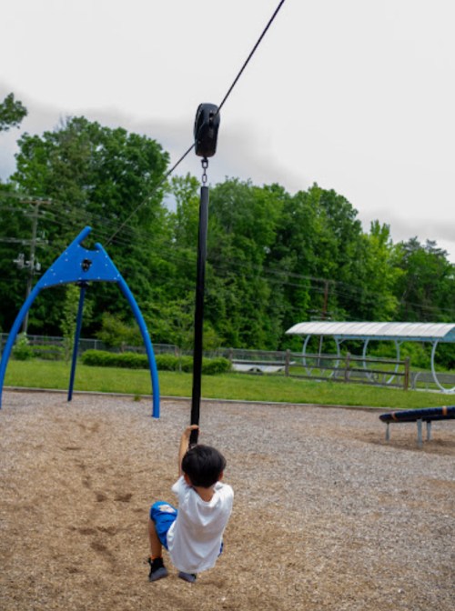 A child swings on a zip line at a playground, surrounded by green trees and a gravel surface.