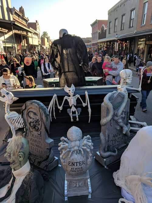 A spooky Halloween parade scene with skeletons, tombstones, and costumed people walking down a festive street.