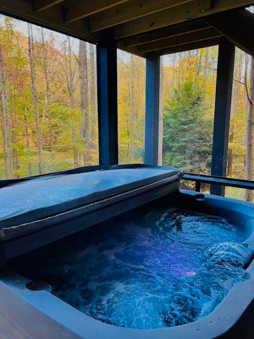 A hot tub with a cover, surrounded by trees and autumn foliage, viewed from a covered porch.