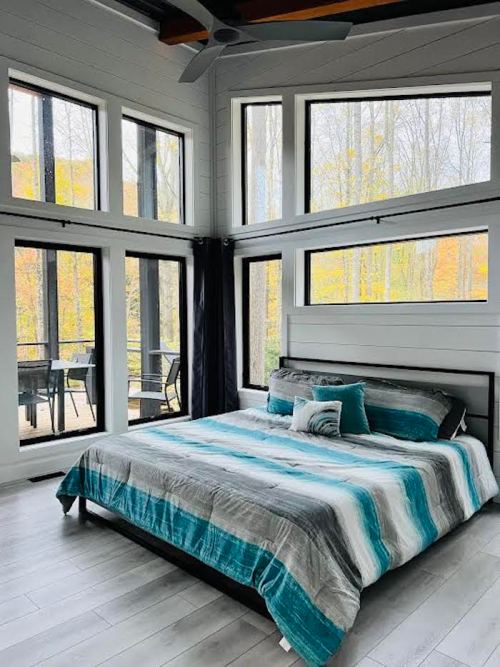 Bright bedroom with large windows, a striped bedspread, and a view of trees outside. Modern and airy design.