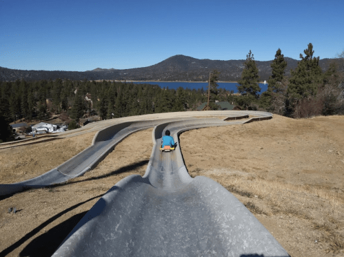 A person rides down a winding slide with a lake and mountains in the background on a clear day.