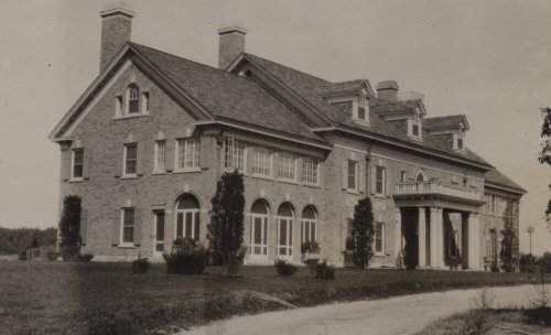 Historic stone mansion with large windows, a portico, and landscaped grounds, set against a clear sky.