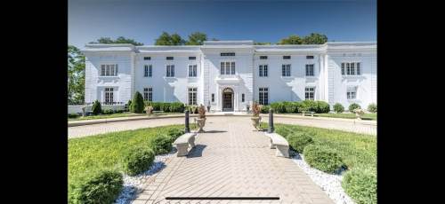 A grand white mansion with a symmetrical facade, surrounded by lush greenery and a paved pathway leading to the entrance.
