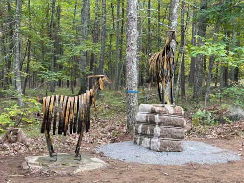 Two metal goat sculptures in a forested area, one standing on a stone base, surrounded by trees and foliage.