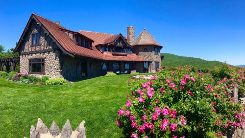 A picturesque stone house with a red roof, surrounded by vibrant flowers and lush green grass under a clear blue sky.