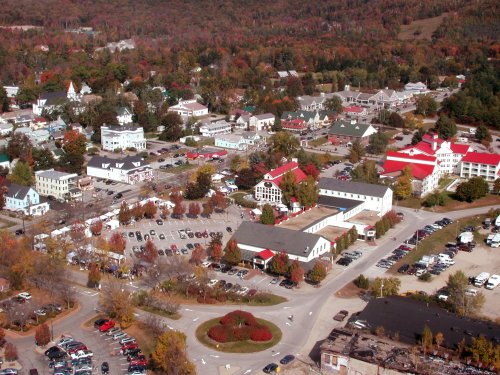 Aerial view of a small town surrounded by colorful autumn foliage, featuring various buildings and parked cars.