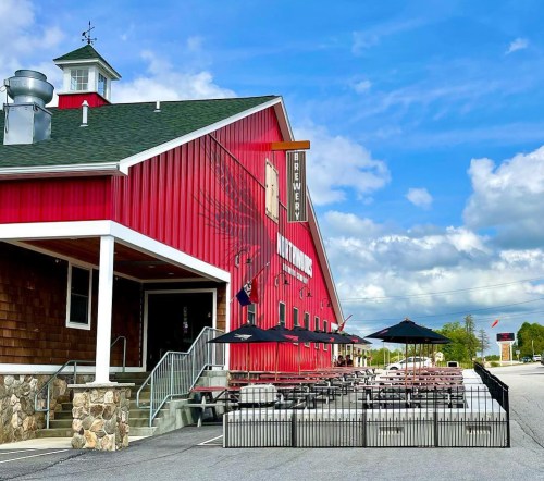 A red brewery building with a stone entrance, outdoor seating, and umbrellas under a blue sky with scattered clouds.